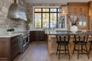 A gorgeous rustic-modern kitchen showcasing the possibilities of funding with a smart kitchen loan, featuring a natural stone accent wall, a light wood island topped with luxurious Cambria Quartz, and a large window overlooking autumn trees.